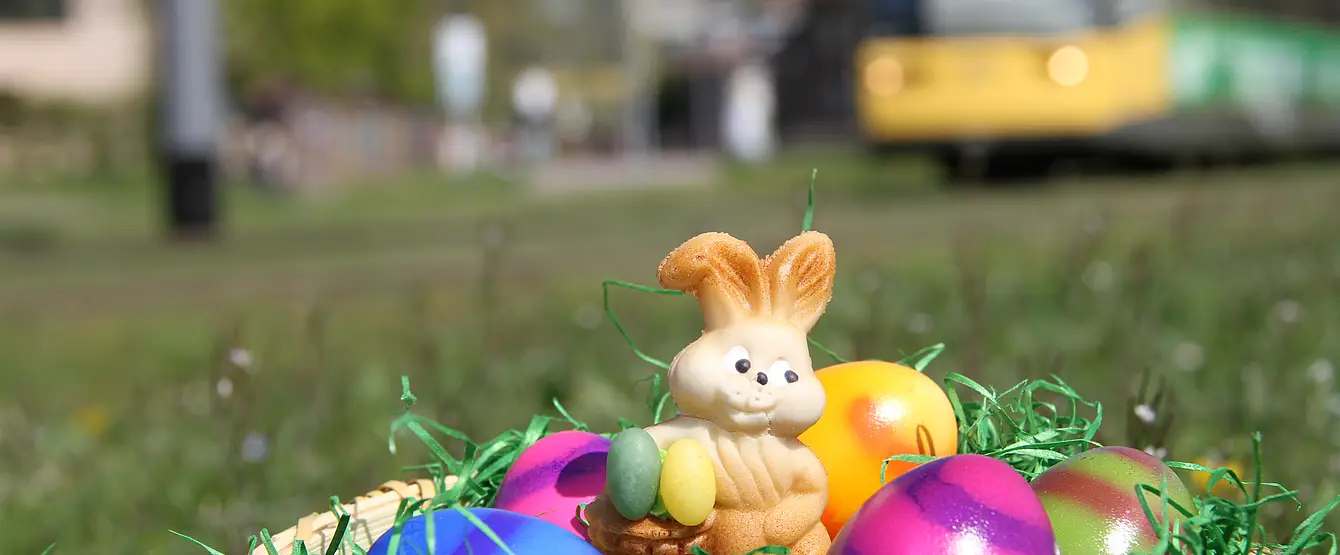 Ein Osternest mit bunten Eisern udn einem Schoko-Hasen liegt auf einer Wiese. Im Hintergrund fährt eine Bahn vorbei.