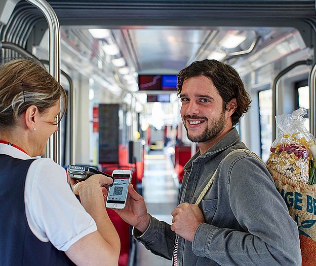Ein Mann mit einer Tasche voller Lebensmitteln über der Schulter zeigt einer Fahrkartenkontrolleurin seine Fahrkarte auf seinem Smartphone.