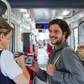 Ein Mann mit einer Tasche voller Lebensmitteln über der Schulter zeigt einer Fahrkartenkontrolleurin seine Fahrkarte auf seinem Smartphone.