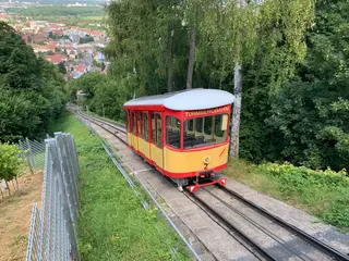 gelb-rotes Fahrzeug der Turmbergbahn auf dem weg zum Gipfel. Rchts sind Bäume und Büsche zu sehen, im Hintergrund die Durlacher Altstadt.