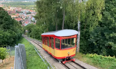 Ein Wagen der Turmbergbahn. Im Hintergrund ist die Durlacher Altstadt zu sehen. Neben der Bahntrasse stehen Bäume.