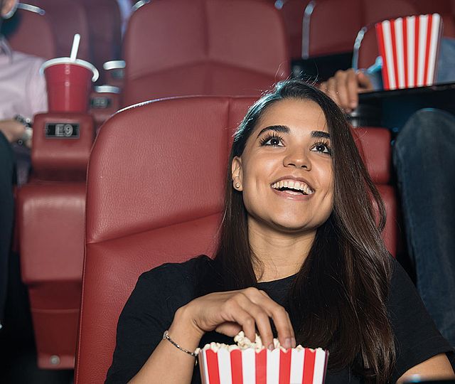 Eine junge Frau sitzt lächelnd in einem Kinosessel und greift in ihre Popcorn-Tüte..