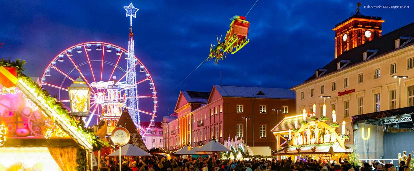 Weihnachtsmarkt Karlsruhe mit beleuchteten Gebäuden, Riesenrad und vielen Besuchern. Der fliegende Weihnachtmarkt fliegt mit seinen Rentieren über den weihnachtlich, geschmückten Platz.