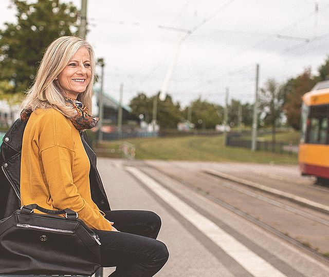 Eine Frau zwischen fünfzig und sechszig Jahren sitzt lächelnd an einer Bahnhaltestelle. Im Hintergrund sieht man eine vorbeifahrende Bahn.