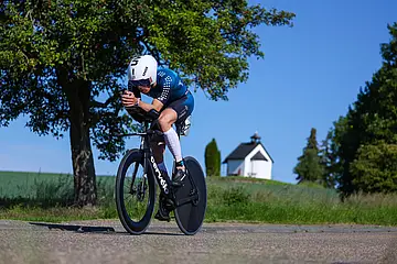 Ein Teilnehmer des Ironman Kraichgau fährt auf einem Rennrad. Im Bildhintergrund sind ein Baum und eine weiße Kapelle zu sehen.