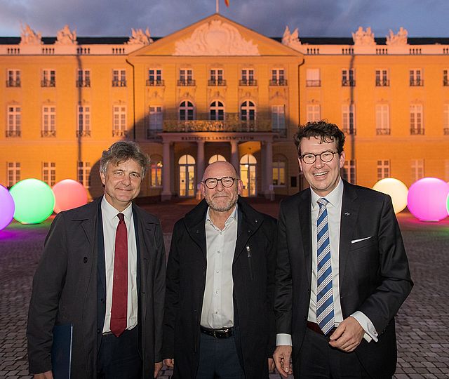 Drei Männer in Anzügen stehen vor dem Karlsruher Schloss