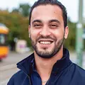 Ein junger Mann steht lächelnd an einer KVV-Haltestelle, im Hintergrund fährt eine KVV-Bahn an ihm vorbei.