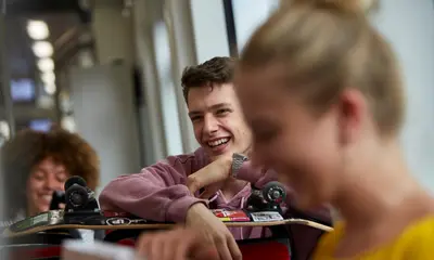 Ein Schüler sitzt in einer Bahn und hält ein Skateboard. Vor und hinter ihm sitzen zwei Mädchen. 