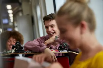 Ein Schüler sitzt in einer Bahn und hält ein Skateboard. Vor und hinter ihm sitzen zwei Mädchen. 