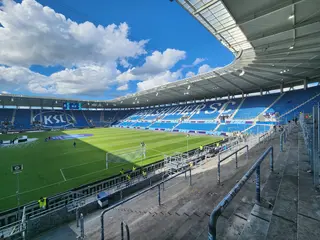 Das Foto zeigt aus der Perspektive der Südtribüne den Stadion-Innenraum im BBBank Wildpark mit Spielfeld und Zuschauertribünen. Weiße Wolken ziehen am blauen Himmel entlang.