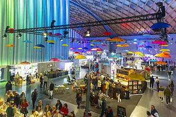 Das Symbolbild zeigt eine der Messehallen während der "offerta" 2025. Zu sehen sind Besucher*innen der Messe und Stände von Ausstellenden. Bunte Regenschirme hängen als Deko an der Hallendecke.