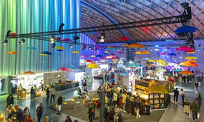 Das Symbolbild zeigt eine der Messehallen während der "offerta" 2025. Zu sehen sind Besucher*innen der Messe und Stände von Ausstellenden. Bunte Regenschirme hängen als Deko an der Hallendecke.