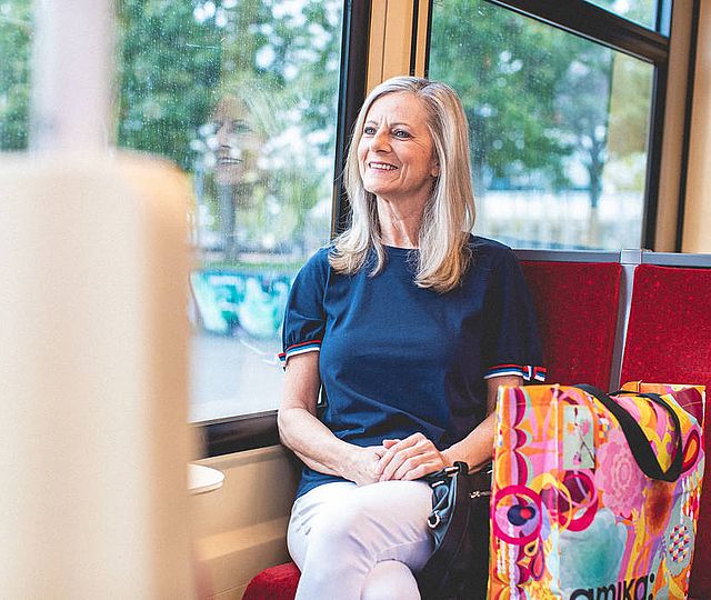 Eine ältere Frau sitzt in der Bahn schaut lächelnd aus dem Fenster., neben ihr steht eine bunte Einkaufstasche.