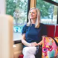 Eine ältere Frau sitzt in der Bahn schaut lächelnd aus dem Fenster., neben ihr steht eine bunte Einkaufstasche.