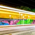Langzeitbelichtung einer Haltestelle bei Nacht. Die Wände der Haltestelle sind bunt bemalt. Man sieht die Licht-Silhouette einer Bahn durchfahren.