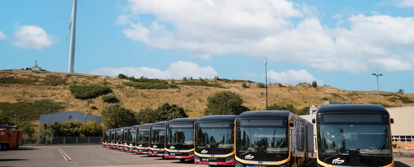 Zwölf neue E-Busse stehen in den VBK-typischen Farben gelb und rot vor einem Windrad.