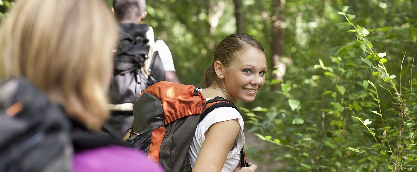 Eine Wandergruppe im Wald. Eine Frau mit Rucksack schaut lächelnd über ihre Schulter nach hinten.