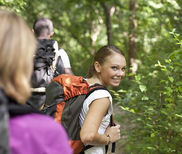 Eine Wandergruppe im Wald. Eine Frau mit Rucksack schaut lächelnd über ihre Schulter nach hinten.