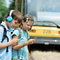 Ein Mädchen und ein Junge mit Schulrucksäcken haben jeweils Kopfhörer auf und schauen auf die Smartphones in ihren Händen. Im Hintergrund ist eine Bahn.
