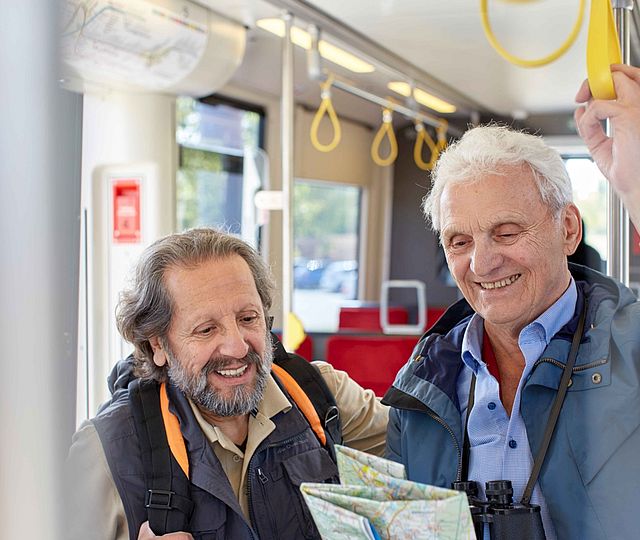 Zwei ältere Herren stehen lachend in der Bahn. Einer trägt einen Rucksack.