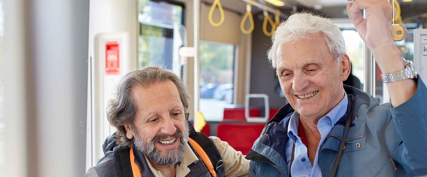 Zwei ältere Herren stehen lachend in der Bahn. Einer trägt einen Rucksack.