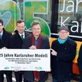 Sechs Männer halten ein Banner. Das Banner bewirbt das Jubiläum 25 Jahre Karlsruher Modell. Hinter den Männern steht eine Bahn, die ebenfalls das Jubiläum bewirbt.