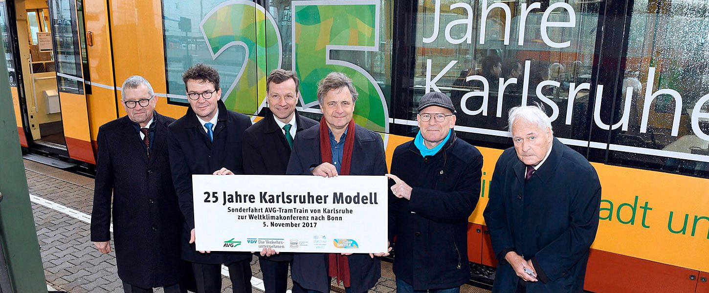 Sechs Männer halten ein Banner. Das Banner bewirbt das Jubiläum 25 Jahre Karlsruher Modell. Hinter den Männern steht eine Bahn, die ebenfalls das Jubiläum bewirbt.