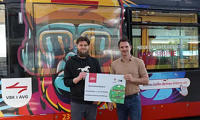 Das Foto zeigt Stefan Mock, Ausbildungsleiter bei VBK und AVG, bei der symbolischen Übergabe eines Deutschlandtickets an Michael aus Bad Herrenalb, der an der Fahrt mit Karaokebahn im April teilgenommen hatte. Beide stehen vor der Karaokebahn im Betriebshof der Verkehrsbetriebe.