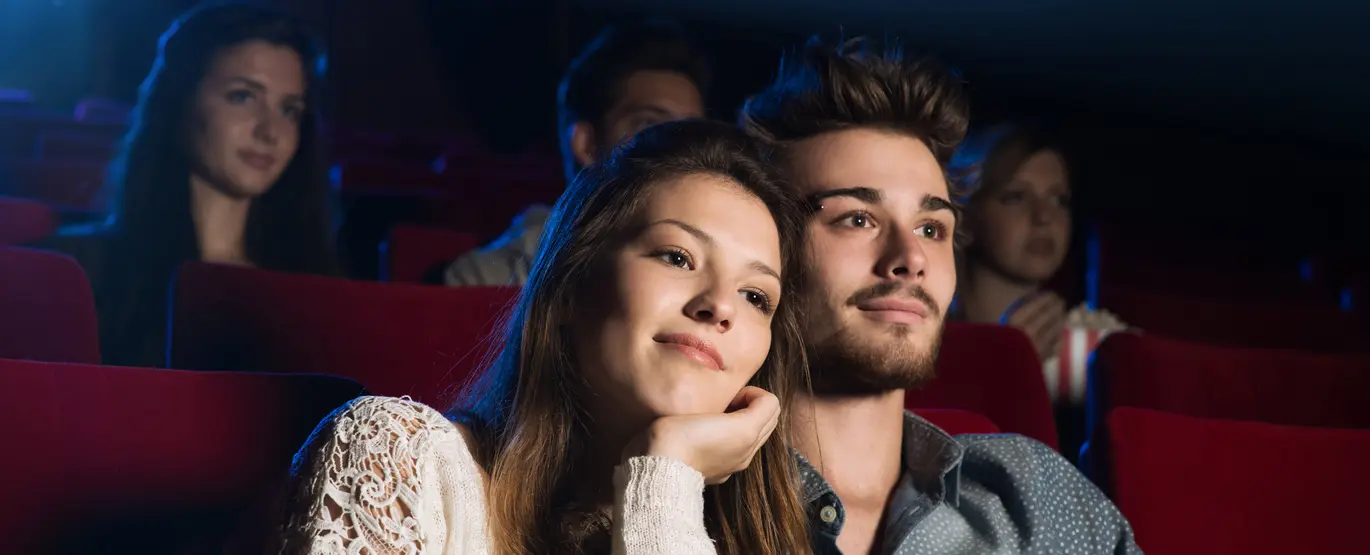 Eine junge Frau und ein junger Mann sitzen im Kino auf Kinosesseln und schauen verträumt Richtung Kino-Leinwand.
