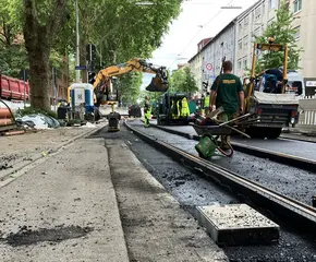 Baustellen-Szenerie mit Bagger und Bauarbeitern. Eine Schubkarre steht links im Vordergrund. 