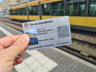 Eine Hand mit dem Deutschlandticket und einer Bahn im Hintergrund