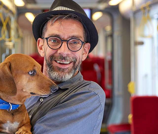 Ein Mann mit Hut und Bart in einer Bahn hält einen Hund auf dem Arm.