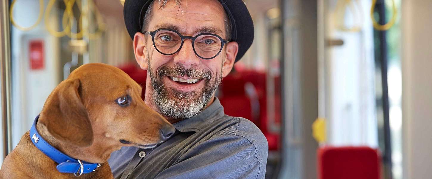 Ein Mann mit Hut und Bart in einer Bahn hält einen Hund auf dem Arm.
