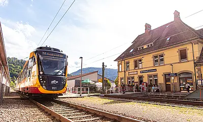 Eine Satdtbahn der AVG steht am Haltepunkt in Forbach. Im Hintergrund ist das historische Bahnhofsgebäude zu sehen.