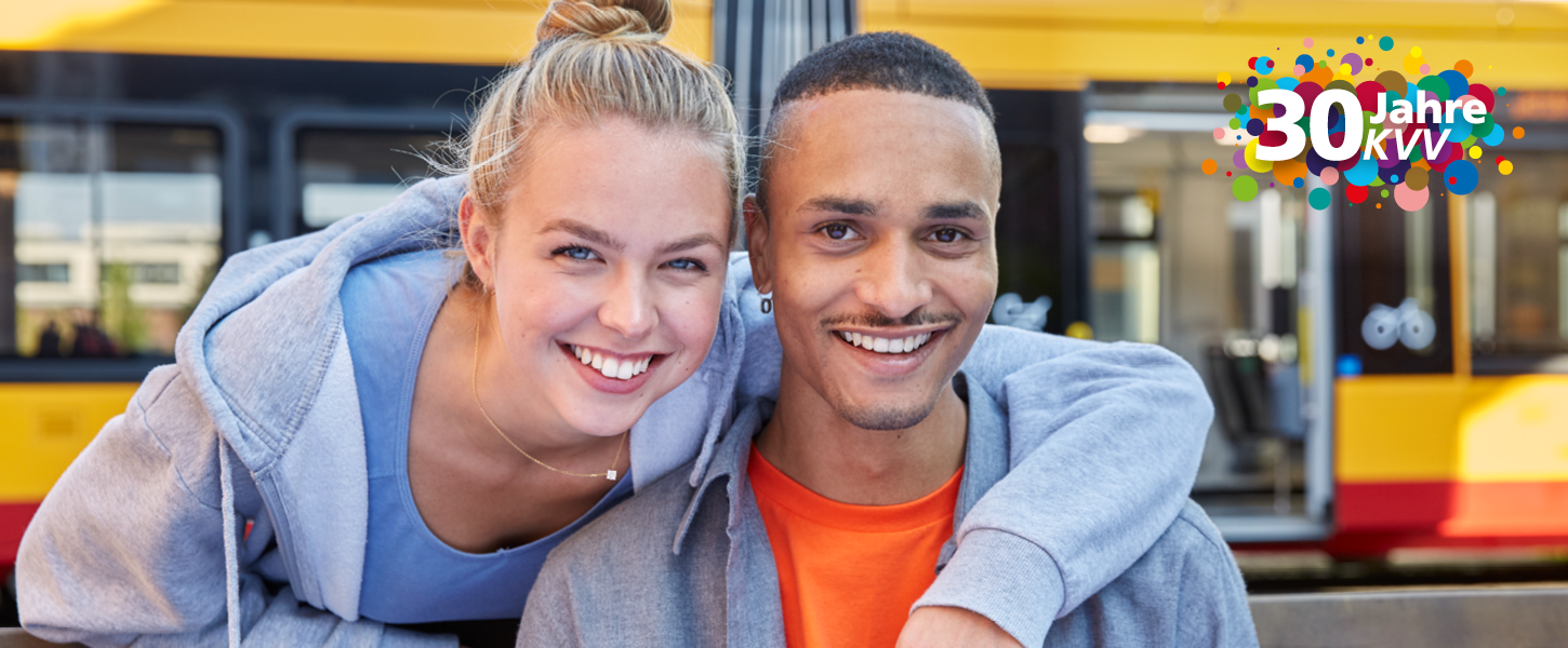 Eine junge Frau hat den linken Arm auf die Schulter eines jungen Mannes gelegt. Beide lächeln freundlich. Im Hintergrund ist eine Bahn zu sehen.
