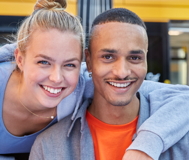 Eine junge Frau hat den linken Arm auf die Schulter eines jungen Mannes gelegt. Beide lächeln freundlich. Im Hintergrund ist eine Bahn zu sehen.