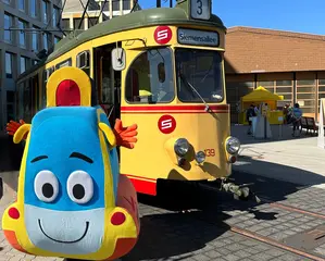 Das Foto zeigt das KVV-Maskottchen Pauly vor einer historischen Straßenbahn