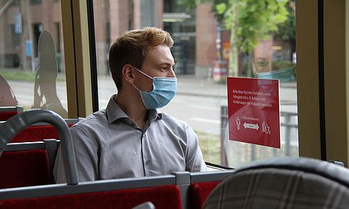Ein männlicher Fahrgast in einer Bahn trägt eine blaue OP-Maske und schaut aus dem Fenster.