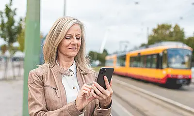 Eine Frau steht in einer Haltestelle und hält ein Smartphone in der Hand. Im Hintergrund ist eine Bahn zu sehen.