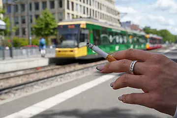Eine Hand hält eine Zigarette an einer Bahnhaltestelle. Im Hintergrund fährt eine Straßenbahn vorbei