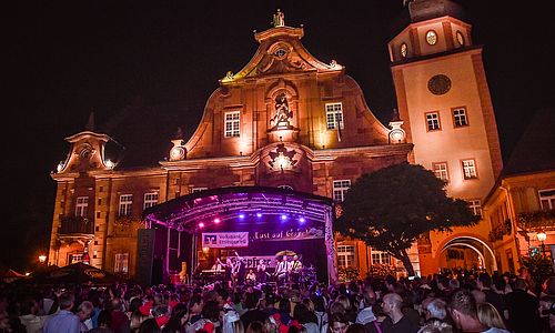 Menschen stehen abends vor einer Musikbühne. Im Hintergrund erhebt sich das beleuchtete Rathaus von Ettlingen.