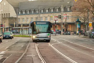 Ein Linienbus fährt auf einer Straße, in der auch Bahngleise liegen. Im Hintergrund ist der Karlsruher Hauptbahnhof zu sehen.
