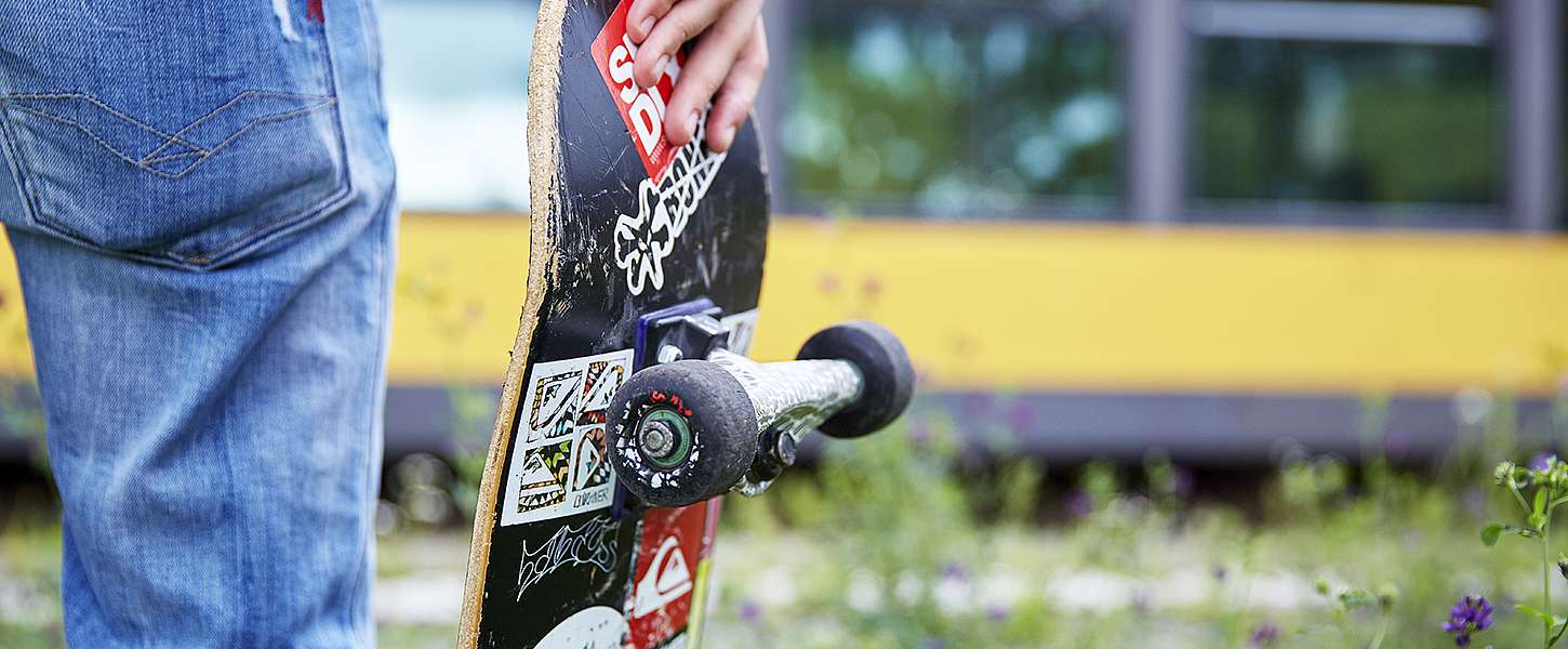 Ein junger Mann, mit der Rückseite zum Betrachter gewandt, hält ein Skateboard in der rechten Hand und schaut vom Gleissteg aus auf die vor ihm stehende gelbe KVV-Bahn.