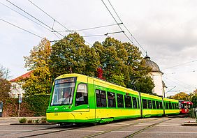 Eine gelb-grüne Niederflurbahn der Albtal-Verkehrs-Gesellschaft auf ihrer Fahrt durch Ettlingen. Im Hintergrund steht ein grüner Baum.