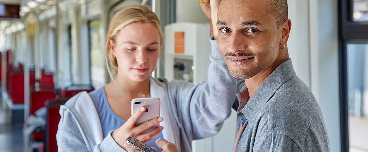 Eine junge Frau und ein junger Mann stehen in einer Bahn. Beide halten ein Smartphone in der Hand.Die Frau schaut auf ihr Smartphone. Der Mann geradeaus und lächelt.