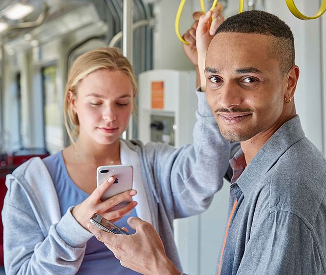 Eine junge Frau und ein junger Mann stehen in einer Bahn. Beide halten ein Smartphone in der Hand.Die Frau schaut auf ihr Smartphone. Der Mann geradeaus und lächelt.