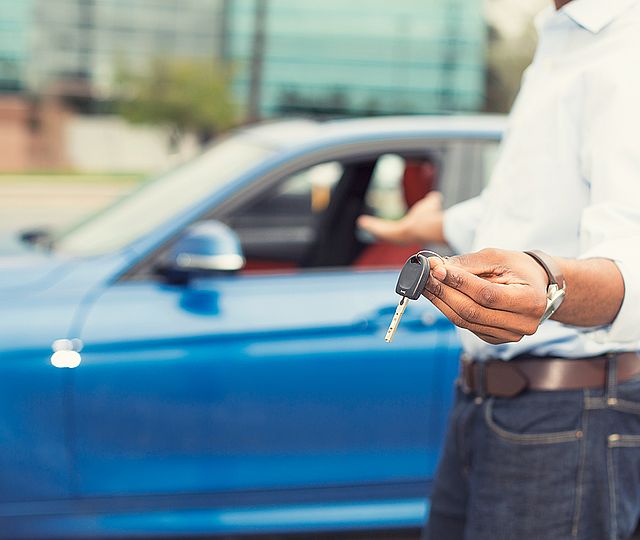 Ein Mann zeigt einen Autoschlüssel und deutet auf ein blaues Auto.