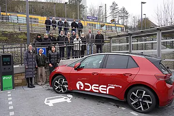 Das Foto zeigt ein rotes Elektroauto des Charsharinganbieters deer vor dem barrierfreien Zugang zur neu gestalteten Stadtbahnhaltestelle. Hinter dem Auto stehen Kämpfelbachs Bürgermeister Thomas Maag (2.von rechts.) und die Projektpartner, Vertreter*innen des Gemeinderats und Bürger*innen der Gemeinde.  