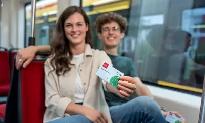 Zwei junge Menschen sitzen in einer Straßenbahn. Eine Frau streckt ihren Arm aus und hält in der Hand das Deutschlandticket