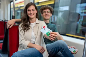 Zwei junge Menschen sitzen in einer Straßenbahn. Eine Frau streckt ihren Arm aus und hält in der Hand das Deutschlandticket
